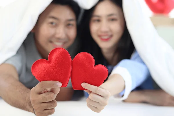 Two red hearts in hand of happy Asian couple lying down on bed together and covering with white blanket. Romantic lover with celebrating anniversary on Valentine Day in decorative bedroom at home.