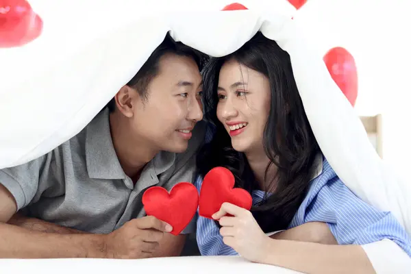 Happy Asian couple holding red heart, looking each other during lying down on bed together. Romantic lover with blanket covering, celebrating anniversary on Valentine Day in decorative bedroom.