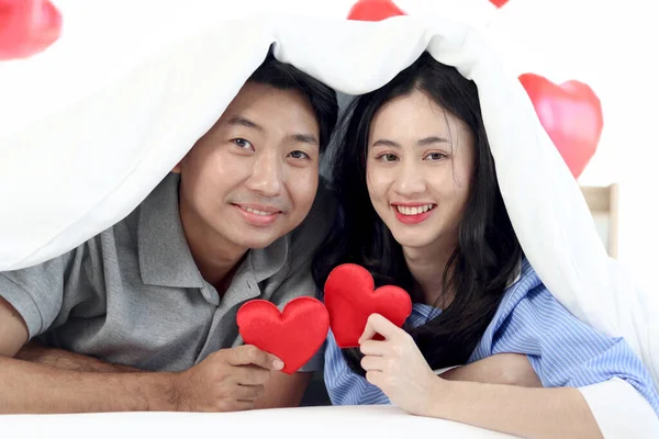Happy cute Asian couple holding red heart during lying down on bed together, covering with white blanket. Romantic lover celebrating anniversary on Valentine Day in beautifully decorative home bedroom