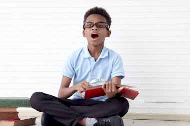 Gözlüklü Afrikalı çocuk beyaz duvar odasında yerde otururken kitap okumak için büyüteç kullanıyor. Kitap yığını, çocuk eğitimi, öğrenme, keşfetme gibi yeni şeylere sahip sevimli bir çocuk..