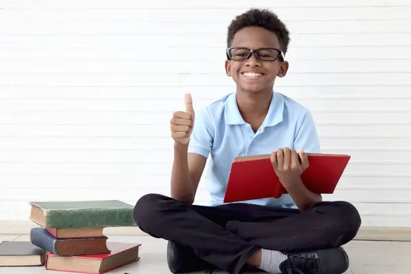 stock image Happy smiling African boy with glasses reading book and giving thumb up while sitting on floor in white wall room. Portrait of cute child with pile, kid education, learning and studying.