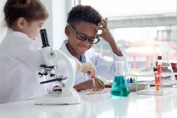 Happy African boy in lab coat doing science experiment with little cute girl buddy friend, young school kid scientist having fun in chemistry laboratory, little children doing research at school.