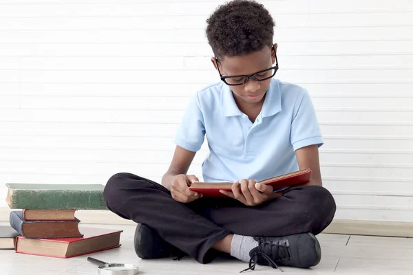 白い壁の部屋の床に座っている間 本を読むために壮大なガラスを使用してアフリカの少年を集中させます 子供の教育 発見を模索するかわいい子供 ストックフォト