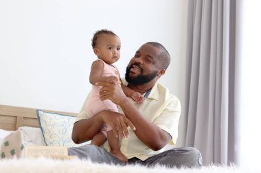 Mutlu Afrikalı bir aile, babası küçük tatlı bebeğini kucağında tutarken beyaz yatakta oturuyor. Evde çocuğuna bakan bir baba, birlikte zaman geçiren bir çocuk..