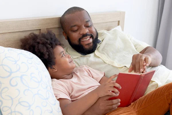 Happy African family, father and son spending time together, young boy with black curly hair and dad reading a book together while lying down on white bed in bedroom. Parent and kid at home.