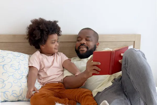 Happy African family, father and son spending time together, young boy with black curly hair and dad reading a book together while lying down on white bed in bedroom. Parent and kid at home.