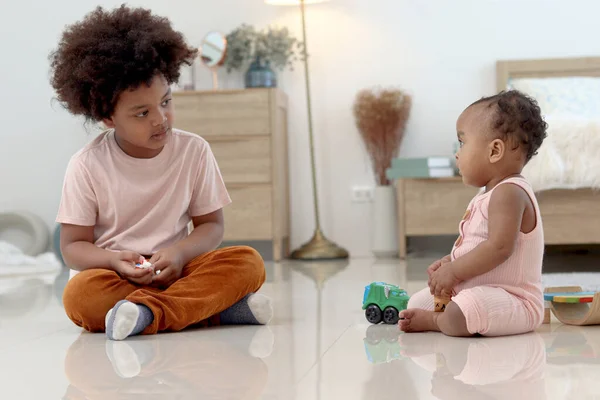 Happy African family spending time together, brother boy with black curly hair sitting on floor, playing toy with little cute toddle baby infant kid in bedroom. Sibling relationships in childhood.