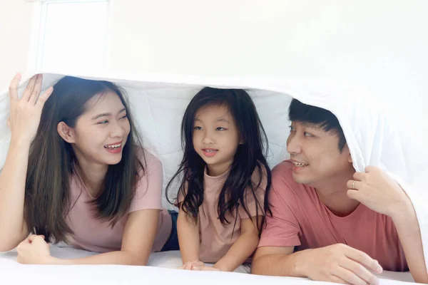 Happy Asian family has fun in bedroom. Father, mother daughter lying in bed together and hiding under white blanket. Parents and kid child girl have good memory at home. Parenthood and childhood