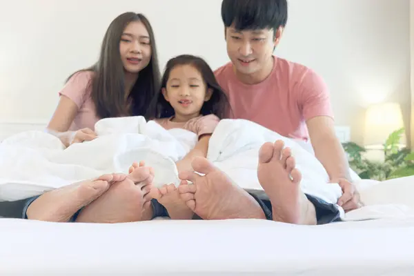 Happy Asian family has fun in bedroom. Feet of mother, father and daughter lying in bed together and hiding under white blanket. Parents and kid girl have good memory at home. Parenthood and childhood
