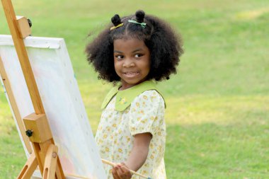 Yeşil bahçedeki tuvale siyah kıvırcık saçlı, tatlı gülümseyen Afrikalı kız. Yeşil yaz parkında doğa resmi çizen bir çocuk. Mutlu küçük çocuk dışarıda eğleniyor..