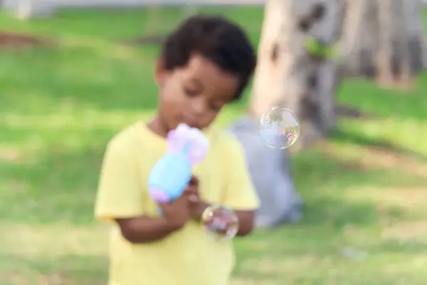 Soap bubble in blued green background, happy African boy making bubbles with blowing bubble gun toy in garden. Kid spends time outdoors in meadow. Child has fun with bubble in summer park