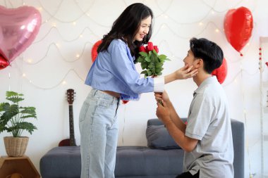 Mutlu Asyalı çift yıldönümlerini birlikte kutluyor. Erkek arkadaşın dizlerine kırmızı gül buketi takacak ve sevgilisine sürpriz yaptığı için evlenme teklif edecek. Sevgililer gününde süslü oturma odasında romantik bir sevgili..
