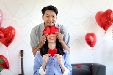 Güzel dekore edilmiş yatak odasında beyaz yatakta otururken gözlerini kapatmak için kırmızı kalp tutan mutlu Asyalı çift, Sevgililer Günü 'nde romantik sevgili, aşk kutlaması.