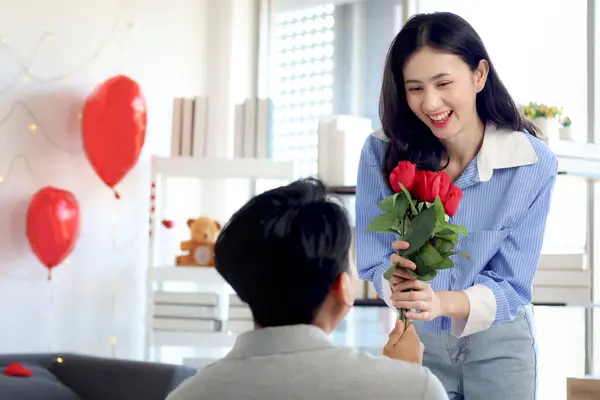 Happy Asian couple celebrating anniversary together. Boyfriend knees to give red rose bouquet and ask girlfriend to marry for surprising her, romantic lover on Valentine Day in decorated living room.