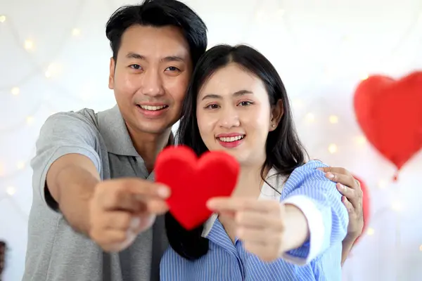Happy cute Asian couple holding and showing red heart to camera while sitting on white bed in beautifully decorated bedroom, romantic lover celebrating anniversary on Valentine Day, love celebration.