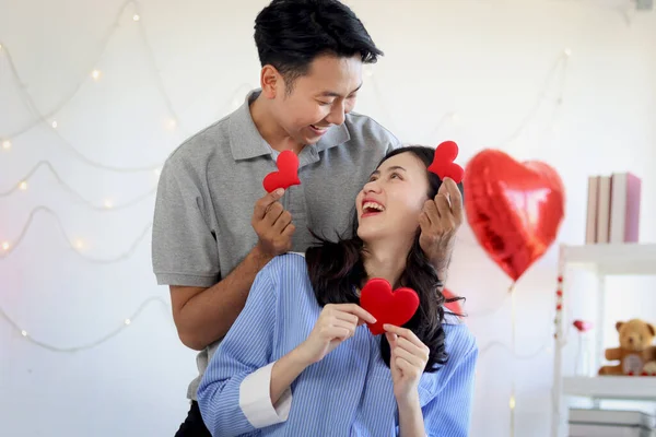 Happy Asian couple holding red heart, looking each other during setting on bed together. Romantic lover celebrating anniversary on Valentine Day in decorative beautiful bedroom. Happy Valentine Day.