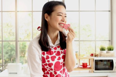 Mutlu gülümseyen, güzel, kırmızı kalp önlüklü, ev yapımı kalp şeklinde kurabiyeler yiyen, mutfak tezgahının arkasında duran, romantik bir sevgililer günü kutlaması hazırlayan Asyalı genç bir kadın..