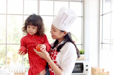 Mutlu aile, gülümseyen güzel Asyalı anne sevimli kırmızı kalp önlüğü takıyor ve küçük kız güzel bir evde elma tutarken şef şapkası kızını tutuyor. Annesi çocuğu taze organik sağlıklı yiyeceklerle besliyor..