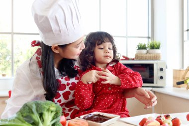 Çikolata yiyen mutlu gülümseyen küçük kız, çikolata kaplı çocuk yüzü. Tatlı kız evdeki mutfakta güzel bir anneyle birlikte yemek pişirmekten hoşlanıyor. Güzel aile anne çocuk konsepti..