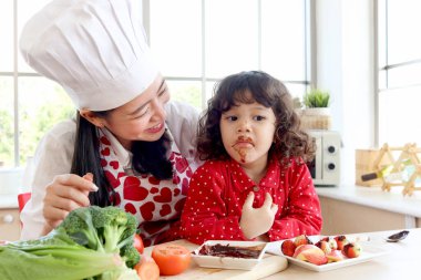 Çikolata yiyen mutlu gülümseyen küçük kız, çikolata kaplı çocuk yüzü. Tatlı kız evdeki mutfakta güzel bir anneyle birlikte yemek pişirmekten hoşlanıyor. Güzel aile anne çocuk konsepti..