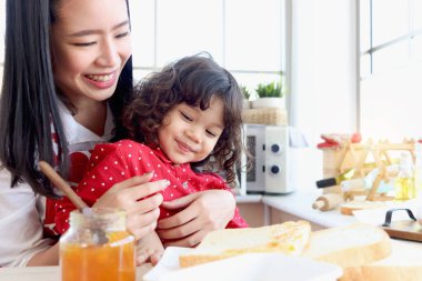 Mutlu aile, gülümseyen tatlı küçük kız, ev mutfağında kızarmış ekmek dilimlerine reçel sürdüğü için annesinin kucağına oturur. Sevimli çocuk sabahları annesiyle kahvaltı etmekten hoşlanıyor..