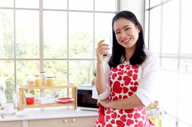 Mutlu gülümseyen, kırmızı kalp önlüklü güzel Asyalı kadın mutfak tezgahının arkasında dururken süt bardağı tutuyor. Kız sabah pencerenin yanında dururken rahatlıyor ve sıcak süt içiyor.