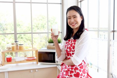 Mutlu gülümseyen, kırmızı kalp önlüklü güzel Asyalı kadın mutfak tezgahının arkasında dururken süt bardağı tutuyor. Kız sabah pencerenin yanında dururken rahatlıyor ve sıcak süt içiyor.