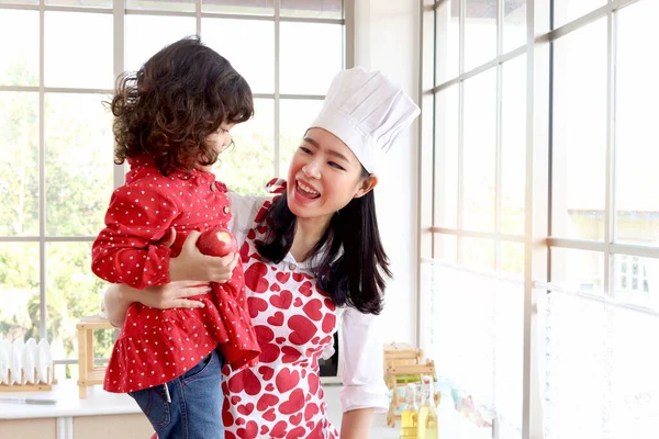 Happy family, smiling beautiful Asian mother wears cute red heart apron and chef hat holds daughter while little girl holds apple in lovely home kitchen. Mom feeds kid with fresh organic healthy food.
