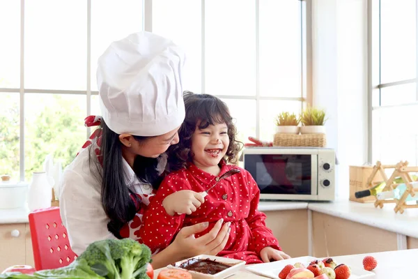 Çikolata yiyen mutlu gülümseyen küçük kız, çikolata kaplı çocuk yüzü. Tatlı kız evdeki mutfakta güzel bir anneyle birlikte yemek pişirmekten hoşlanıyor. Güzel aile anne çocuk konsepti..