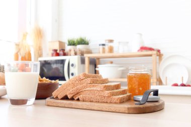 Tam tahıllı ekmek sırası reçel ve bir bardak sütle ahşap masa üzerinde bulanık mutfak arka planı. Sabahları basit bir kahvaltı hazırlamak için gerekli malzemeler. Proteinli, kalsiyumlu ve vitaminli yiyecekler.