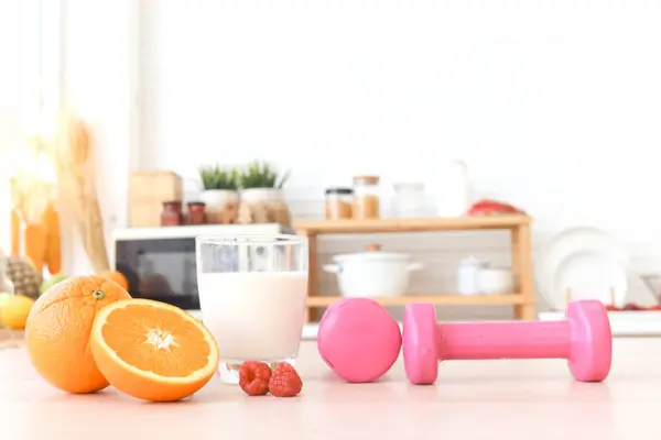 Dumbbells, orange fruit, raspberry, milk on wooden table with blur cozy kitchen background. Healthy food and exercises for losing weight. Food with protein, calcium, vitamins rich and fitness workout