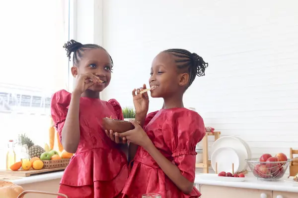 African twin girl sister with curly hair braid African hairstyle eating snack chips in kitchen. Happy smiling kid sibling spending time together. Cute children in lovely family.