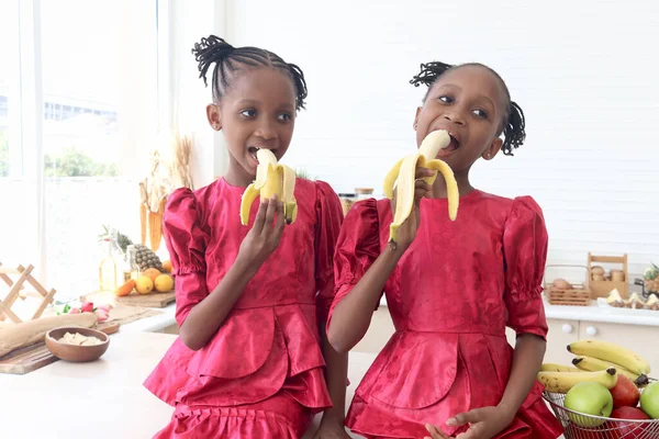 African twin girl sister with curly hair braid African hairstyle eating banana in kitchen. Happy smiling kid sibling eating fruit and spending time together. Cute children in lovely family.