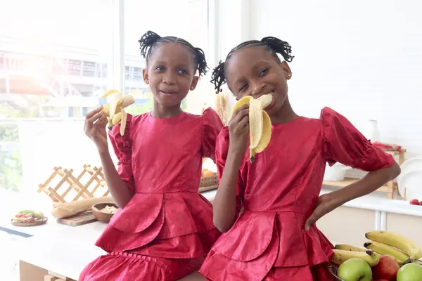 African twin girl sister with curly hair braid African hairstyle eating banana in kitchen. Happy smiling kid sibling eating fruit and spending time together. Cute children in lovely family.
