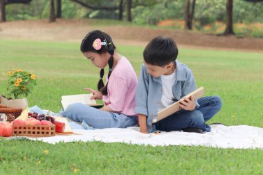 Mutlu çocuklar yaz yeşil park bahçesinde piknik yapıyorlar. İki Asyalı arkadaş, erkek ve kız kitap okuyor, sırt sırta verip bir paspasa oturuyorlar. Çocuklar dışarıda birlikte oynuyorlar. Neşeli çocuklar açık havada olmayı sever.