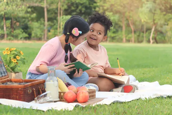 Happy children have a picnic in summer park, cute curly hair African girl with Asian buddy friend studying, reading book together while sitting on mat on green grass. Kid do homework in outdoor garden