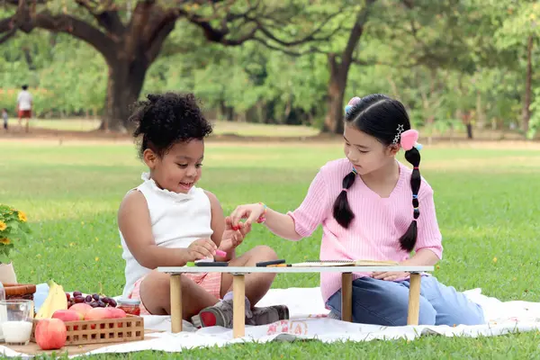 Happy children having a picnic in summer park, cute curly hair African girl with Asian buddy friend studying while sitting on mat on green grass. Kid doing homework together in outdoor garden.