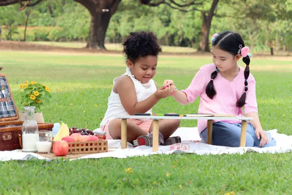 Happy children having a picnic in summer park, cute curly hair African girl with Asian buddy friend studying while sitting on mat on green grass. Kid doing homework together in outdoor garden.