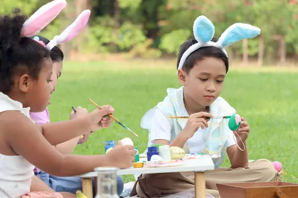 Happy three children in park, cute Asian boy with his friends wearing bunny ears, painting  Easter eggs with paintbrush together on green grass in garden. Kids celebrate Easter holiday outdoor.