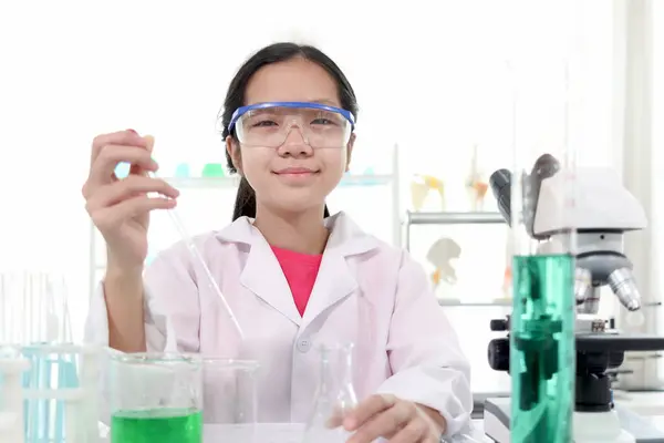 Cute young scientist schoolgirl wearing lab coat and safety glasses, doing science experiments. Portrait of happy student girl child studying chemistry in laboratory. Kid learning science education.