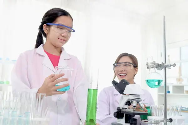 Cute young scientist schoolgirl wears lab coat and safety glasses, doing science experiments under guidance of teacher. Student girl child study chemistry in laboratory. Kid learning science education