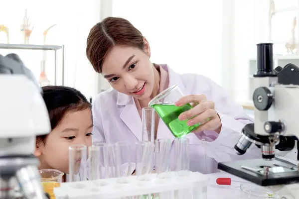 Beautiful Asian female teacher in lab coats teaching schoolgirl child to do science experiments. Happy young scientist kid girl using lab equipment for research study in school laboratory.