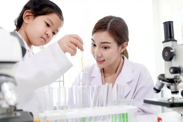 Beautiful Asian female teacher in lab coats teaching schoolgirl child to do science experiments. Happy young scientist kid girl using lab equipment for research study in school laboratory.