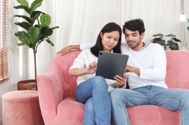 Oturma odasında mutlu bir çift, Asyalı bir kadın erkek arkadaşıyla kanepede oturuyor, evde birlikte zaman geçirirken tablet kullanıyorlar. Aşık olmak ve evde romantik anlar yaşamak.