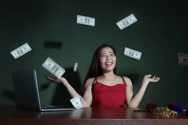 Happy young Asian woman in red sexy dresses costume working online on laptop computer and throwing money banknotes (mockup) to success celebration. Rich crazy funny winner throwing out money.