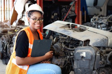 Kıvırcık saçlı, güvenlik yeleği giyen Afro-Amerikalı bir kadın, elinde belge tutarken yedek parça deposundaki ürün stoklarını incelerken sayısız ikinci el motor parçalarıyla çevrili..
