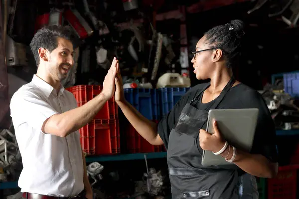 Happy harmony people work together at workplace, African American woman making high five hand touch with colleague at auto spare parts store warehouse, surrounded by secondhand engine parts.