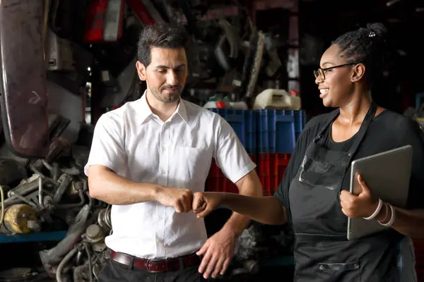 Happy harmony people work together at workplace, African American woman fist bumping hand after complete work with colleague at auto spare parts store warehouse, surrounded by secondhand engine parts.