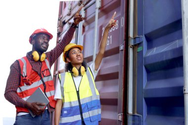 Güvenlik yeleği ve kask giyen iki Afrikalı mühendis ve kadın lojistik kargo konteynerinde birlikte çalışarak konteynırı işaret ediyorlar. İş yerinde çalışanlar.