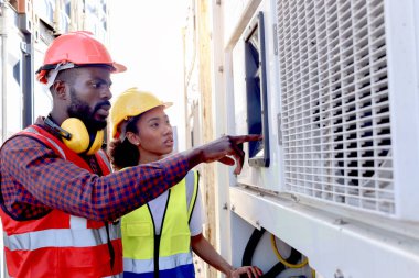 İki endüstriyel Afrikalı mühendis erkek ve kadın güvenlik yeleği ve miğfer giyer, imalat fabrikasında veya lojistik kargo deposunda kontrol düğmesi takarlar. İş yerinde çalışanlar